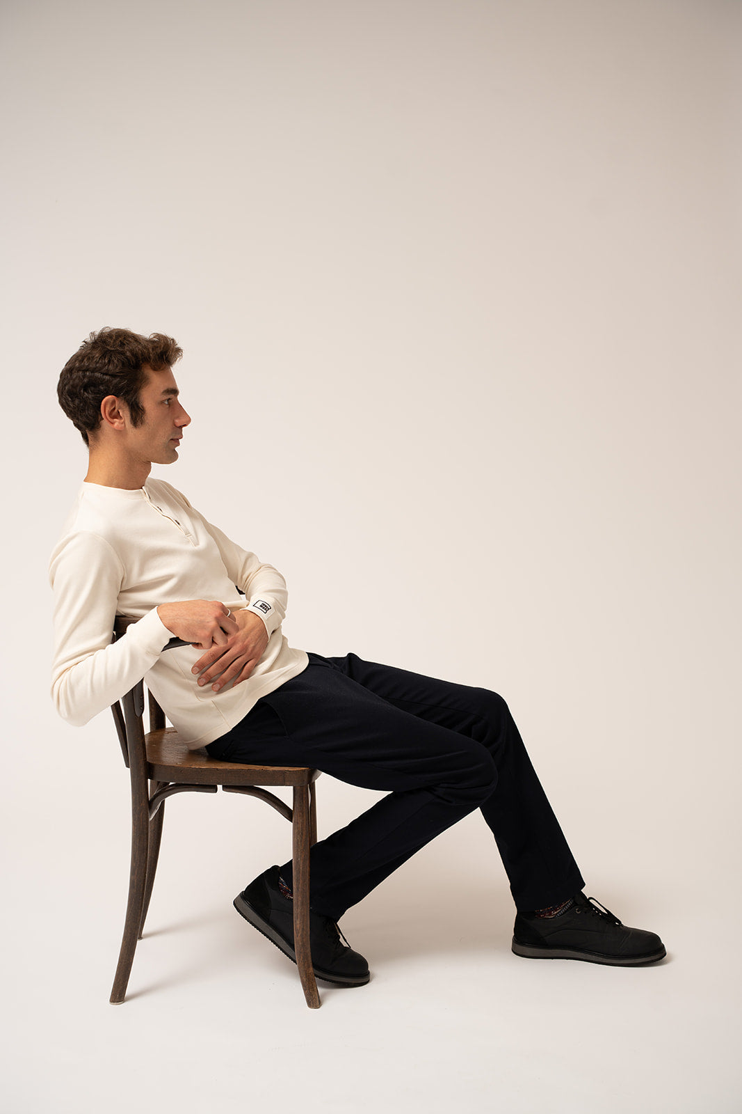A man sitting on a chair wearing a white shirt and navy blue chino pants against a plain background